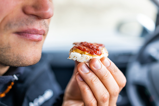 Macro closeup of man hand holding one rice cake by mouth in car road trip blurry background topped with peanut butter, strawberry jam as vegan dessert snack