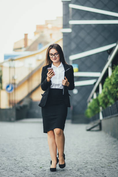 istock Young beautiful business woman walks next to an office building with documents and using the phone while distracted from work. Horizontal photo 1135625544