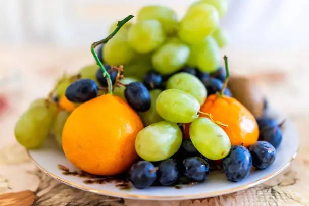 Closeup of whole mandarin satsuma orange and green grapes fruits with blue red grapes pile heap on plate bowl in winter autumn season