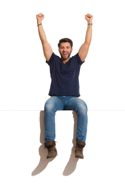 Young man in boots, jeans and blue shirt is sitting on a top, rising arms, looking at camera and shouting. Full length studio shot isolated on white.