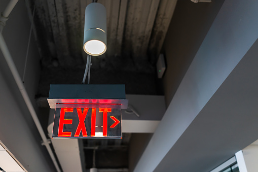 Red exit sign hanging from ceiling looking up view with arrow direction and color illuminated fixture in office building closeup