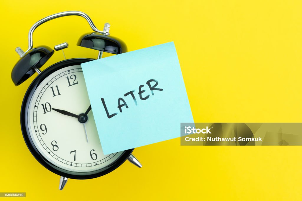 Sticky post with handwriting the word Later stick on alarm clock on solid yellow background with copy space using as procrastination, self discipline or laziness concept Sticky post with handwriting the word Later stick on alarm clock on solid yellow background with copy space using as procrastination, self discipline or laziness concept. Wasting Time Stock Photo