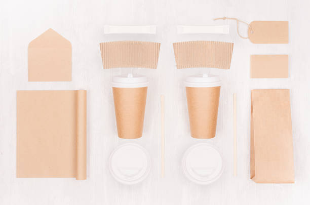 Coffee takeaway set mockup for brand - two brown paper cups with blank notebook, card, label, packet, sugar, cap, envelope on white wood board, top view.