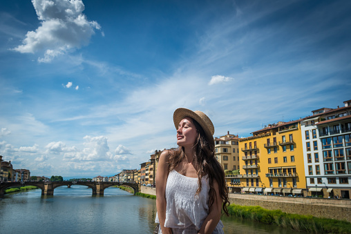 Young woman enjoys holidays in Florence, Italy. Beautiful girl with smile and closed eyes in old town sits with view on river, sky, houses and bridge.