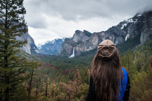 Young female traveler standing back and looking on forest, mountains, waterfall and trees. Girl on background of amazing valley in Yosemite park, USA.