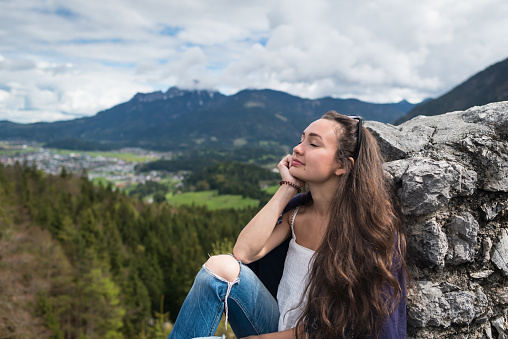 Female traveler sits on peak of mountain with closed eyes on background of valley, forest and village. Young Kazakh girl enjoying day in Alps, Austria
