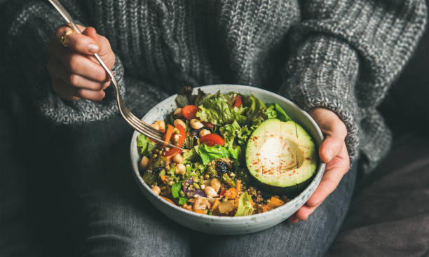 Woman eating healthy vegetarian dinner from Buddha bowl, close-up Healthy vegetarian dinner. Woman in jeans and warm sweater holding bowl with fresh salad, avocado, grains, beans, roasted vegetables, close-up. Superfood, clean eating, vegan, dieting food concept foxys_forest_manufacture stock pictures, royalty-free photos & images