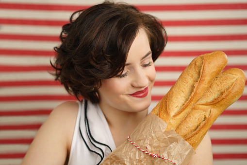 Pretty young woman in a summer sleeveless blouse holding freshly baked baguettes in a brown paper packet over a striped red and white blurred background