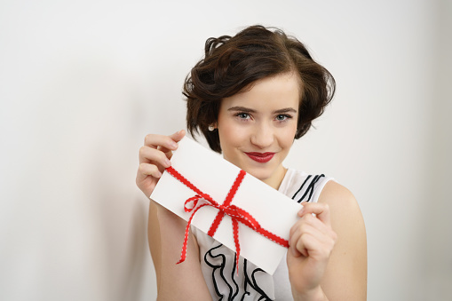 Young woman holding up a gift card tied with a romantic red bow and ribbon with a smile as she leans against an interior white wall