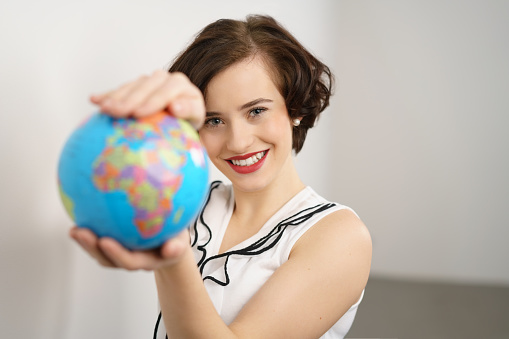 Pretty stylish young woman holding a world globe extended to the camera in a concept of travel or saving the planet, with focus to her face