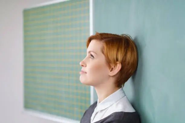 Side portrait of young red haired woman with short haircut leaning back on green chalkboard and looking up, standing in classroom. Copy space