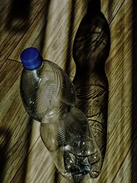 Abstract view of empty clear plastic bottle with water vapor on the bottle wall and strong shadow on oak textured wooden floor in strong day light. Selective focus. Fine art and abstract concept. Recycling issue concept.