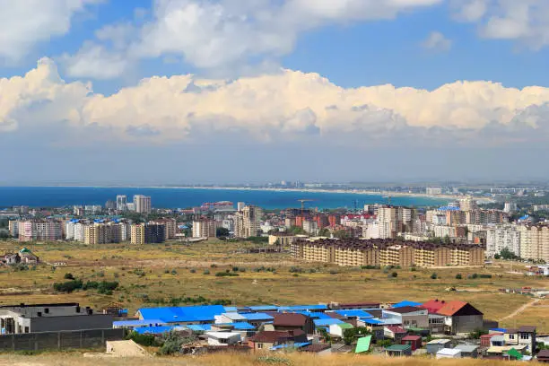 Aerial panorama of Anapa resort city, Russia. Summer day under blue cloudy sky. View from mountain. New buildings, construction cranes and Black Sea water.