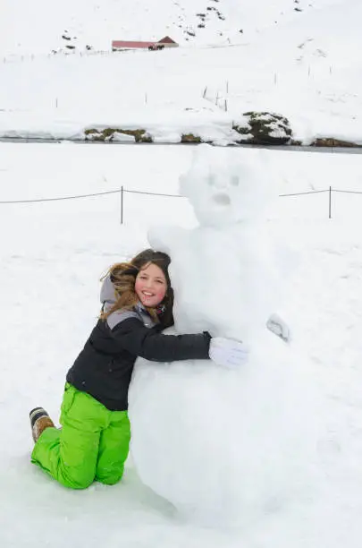 Girl proud of the snowman she built, she is hugging it and has a huge smile on her face.
