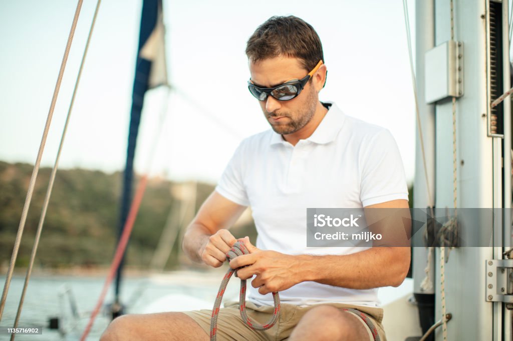 Skipper of the sailboat Handsome mid adult skipper on sailboat fixing and examining ropes and tying knots while sitting on boat deck Tying Stock Photo