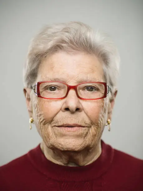 Close up portrait of senior caucasian woman with blank expression against gray white background. Vertical shot of spanish real people staring in studio with white hair and glasses. Photography from a DSLR camera. Sharp focus on eyes.