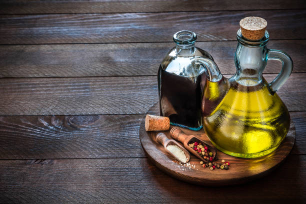 High angle view of two bottles full of olive oil and balsamic vinegar over dark brown cutting board on rustic wooden table. In front of the bottles are two serving scoops with salt and pepper. Copy space at left part of the image. predominant colors are brown and yellow. Low key DSLR photo taken with Canon EOS 6D Mark II and Canon EF 24-105 mm f/4L