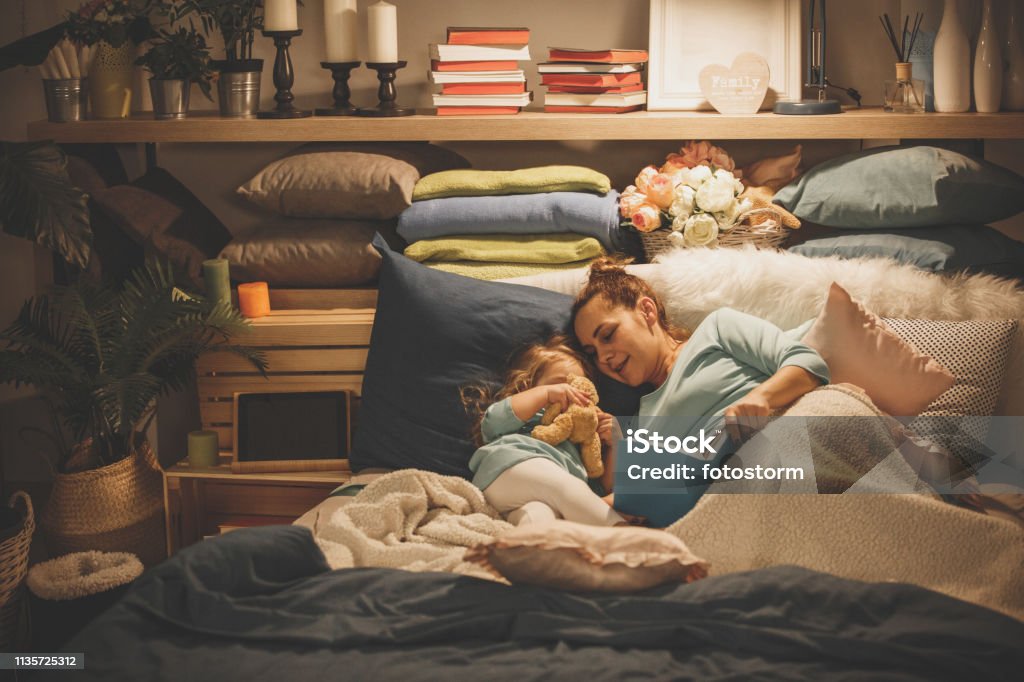 Mother reading a book to her daughter Mother is reading a book before bedtime while her sleepy daughter is lying down next to her. Storytelling Stock Photo