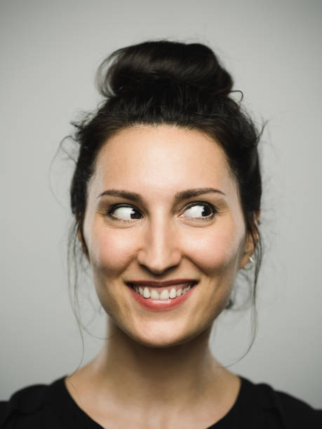 Studio portrait of real mediterranean young woman with excited expression looking to the side Close up portrait of mediterranean young woman with excited expression looking to the side against gray white background. Vertical shot of caucasian real people laughing in studio with long black hair and a hair bun. Photography from a DSLR camera. Sharp focus on eyes. sideways glance stock pictures, royalty-free photos & images