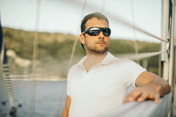 Handsome mid adult skipper on sailboat leaning on sail boom. He wears white t-shirt and sunglasses.