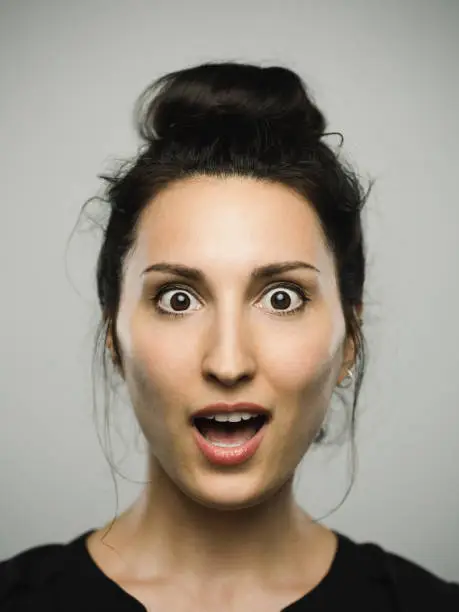 Close up portrait of mediterranean young woman with surprised expression against gray white background. Vertical shot of caucasian real people shocked in studio with long black hair and a hair bun. Photography from a DSLR camera. Sharp focus on eyes.