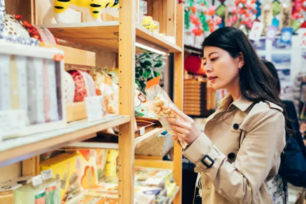 young japanese mom choosing snack for kids in local specialty shop in dotonbori osaka japan. asian mother buying food cookies for family after work in the vendor. beautiful lady reading the mark.