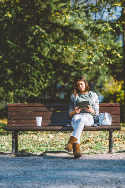 istock Full Length Of Young Woman Using Digital Tablet While Sitting On Bench Against Trees In Park 1135757777