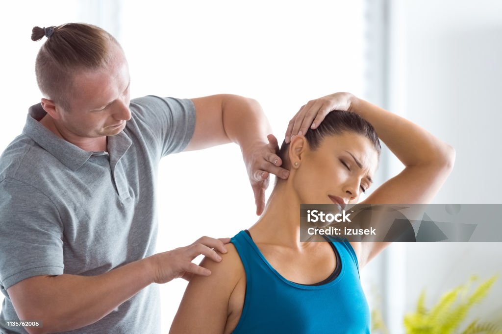 Physiotherapist massaging young woman's neck Physical therapist giving neck massage to young woman in his office. Patient sitting on massage table. Chiropractic Adjustment Stock Photo