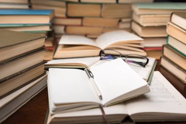 istock Education learning concept with open book and notebook on the table. Stack piles of books on reading desk and glasses in bookshelves background 1135763978