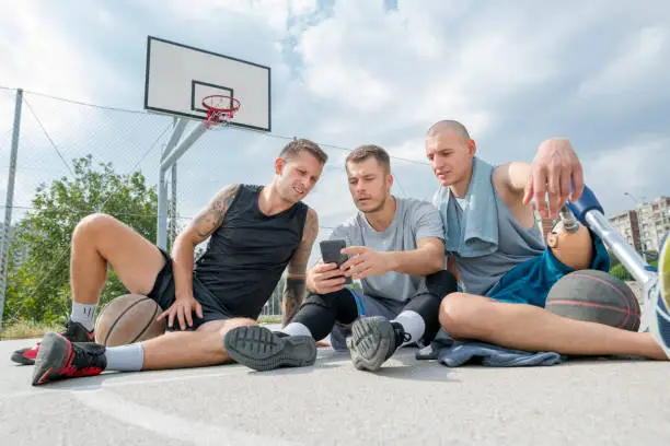 Young man with amputated leg and his friends sitting at the outdoor courtyard, cooling down after a basketball training and using a smart phone.