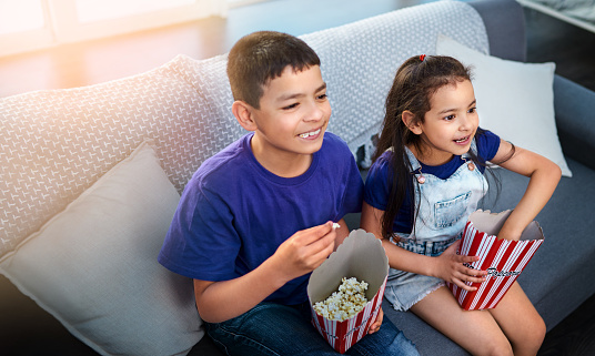 High angle shot of two young children sitting on a sofa and eating popcorn while watching movies at home