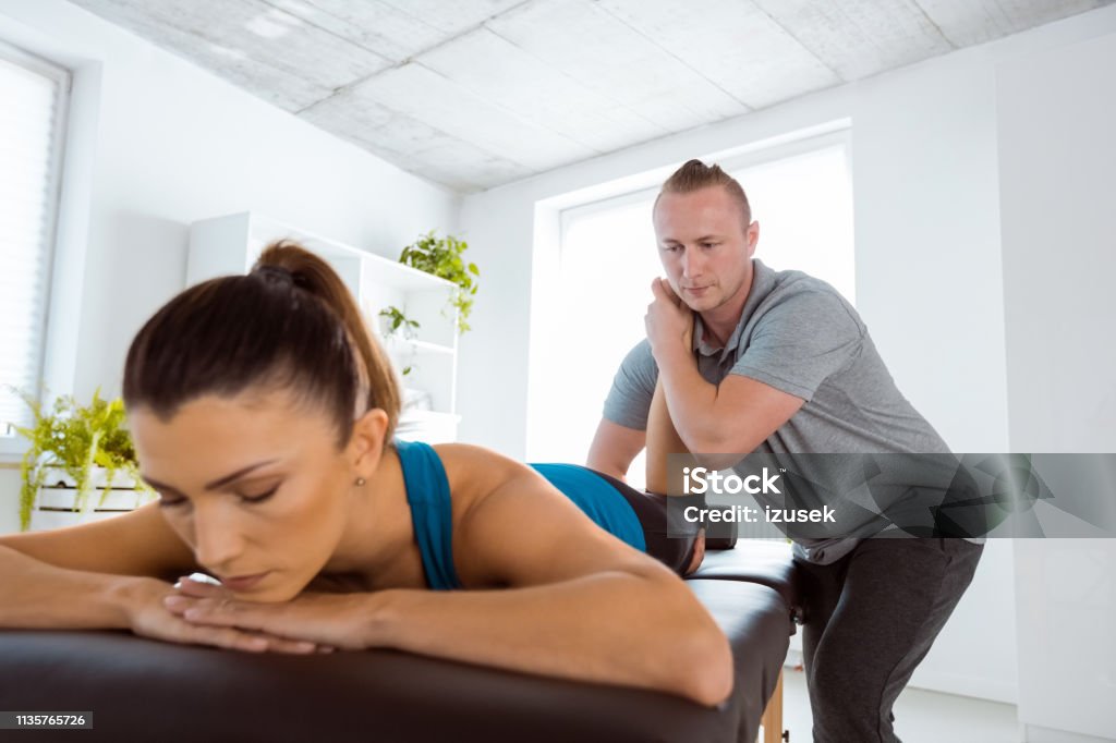 Massage therapist stretching young woman's legs Physical therapist giving legs massage to young woman in his office. Patient lying on massage table. Adult Stock Photo