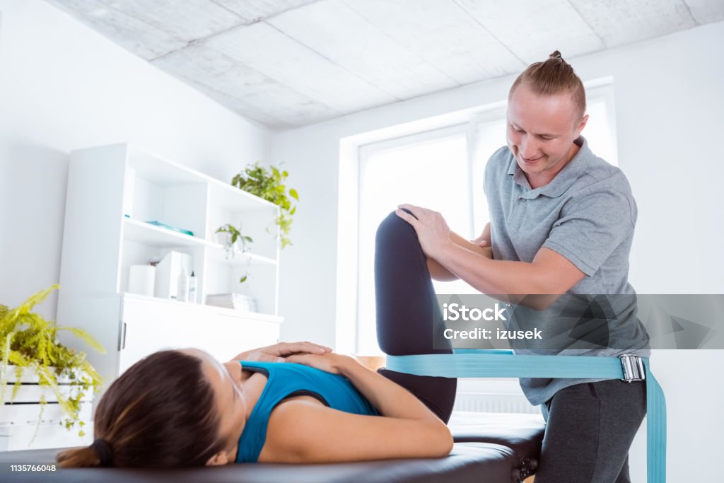 Physiotherapist massaging young woman Physical therapist giving leg massage to young woman, using traction belt. Patient lying down on massage table. Physical Therapy Stock Photo