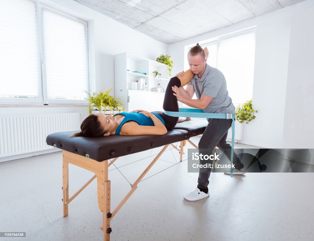 Physiotherapist massaging young woman Physical therapist giving leg massage to young woman, using traction belt. Patient lying down on massage table. Physical Therapy Stock Photo