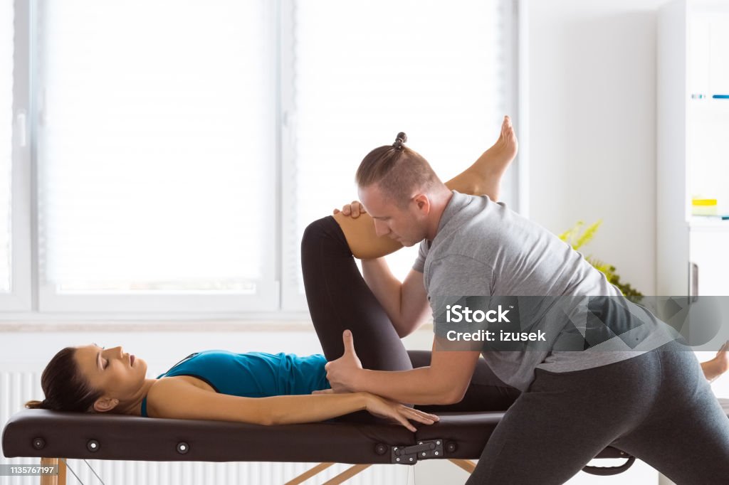 Physiotherapist stretching young woman's legs Physical therapist giving legs massage to young woman in his office. Patient lying on massage table. Adult Stock Photo