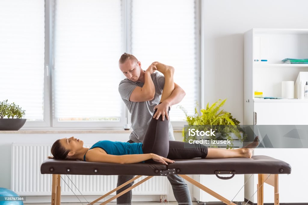 Physiotherapist stretching young woman's legs Physical therapist giving legs massage to young woman in his office. Patient lying on massage table. Physical Therapy Stock Photo