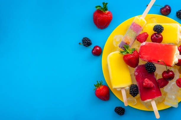Summer treats and sweet frozen desserts concept with assorted popsicles and berry fruits like strawberry, cherry, blackberry and raspberry isolated on minimalist blue background with copyspace