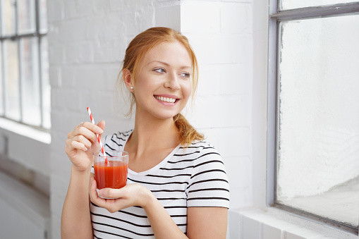 Single happy young red haired woman holding carrot juice with red and white straw near window