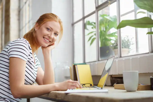 Smiling happy young woman studying at home sitting at a work table with her notes and laptop looking at the camera with a friendly grin