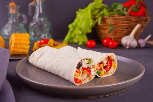 Burrito rolls with vegetables on black table and vegetables, cherry tomatoes and garlic at background