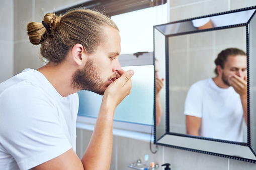 Cropped shot of a young man smelling his breath while standing in his bathroom