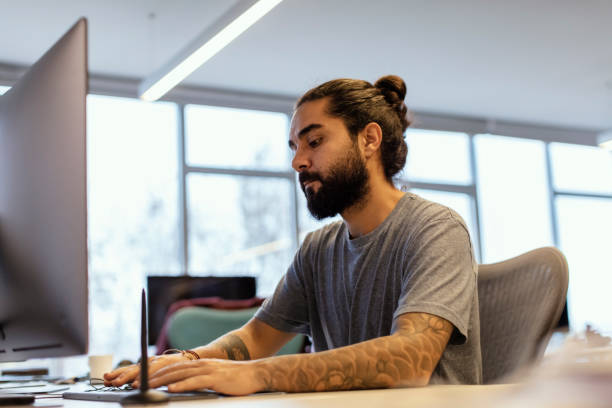 istock Portrait of Stylish Hipster Guy with  Tattoo on Hand, Writes Notes in Computer at Office 1135788256