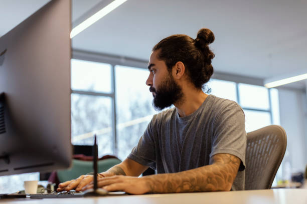 istock Portrait of Stylish Hipster Guy with  Tattoo on Hand, Writes Notes in Computer at Office 1135793236