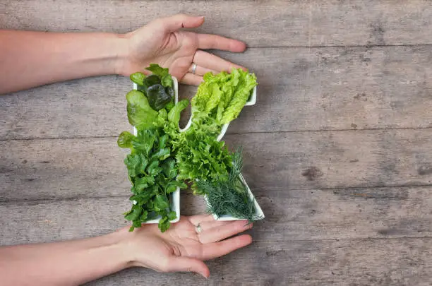 Vitamin K nutrient in food concept. Woman's hands holding letter K shaped plate with different fresh leafy green vegetables, herbs, lettuce on wooden background. Flat lay or top view.