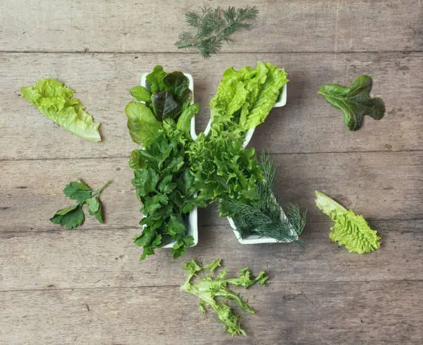 Vitamin K in food concept. Plate in the shape of the letter K with different fresh leafy green vegetables. Some   lettuce and herbs lie near on wooden background. Flat lay or top view.