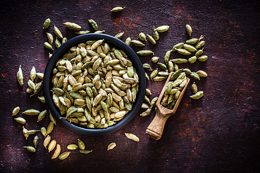 Spices: Top view of a black bowl filled with cardamom pods shot on abstract brown rustic table. A wooden serving scoop is beside the bowl and cardamom pods are scattered on the table. Useful copy space available for text and/or logo. Predominant colors are green and brown. Low key DSRL studio photo taken with Canon EOS 5D Mk II and Canon EF 100mm f/2.8L Macro IS USM.