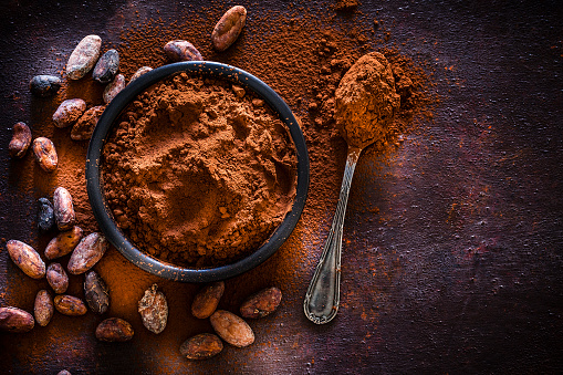 Top view of a black bowl filled with cocoa powder shot on abstract brown rustic table. A metal spoon with cocoa powder is beside the bowl and some cocoa beans are scattered on the table. Useful copy space available for text and/or logo. Predominant colors is brown. Low key DSRL studio photo taken with Canon EOS 5D Mk II and Canon EF 100mm f/2.8L Macro IS USM.