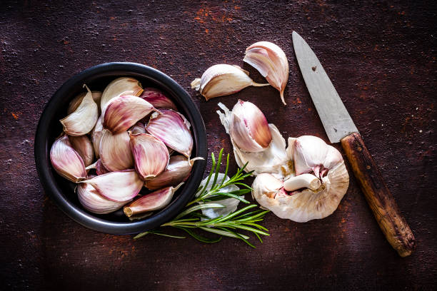 Garlic bulb and cloves shot from above on rustic brown background Vegetables: Garlic cloves in a black bowl shot from above on rustic brown background. A garlic bulb an three cloves are beside the bowl. An old kitchen knife is at the right of an horizontal frame. Predominant colors are brown and white. Low key DSRL studio photo taken with Canon EOS 5D Mk II and Canon EF 100mm f/2.8L Macro IS USM. garlic stock pictures, royalty-free photos & images