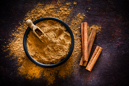 Spices: Top view of a black bowl filled with ground cinnamon shot on abstract brown rustic table. Cinnamon sticks are beside the bowl and ground cinnamon is scattered on the table. Useful copy space available for text and/or logo. Predominant colors is brown. Low key DSRL studio photo taken with Canon EOS 5D Mk II and Canon EF 100mm f/2.8L Macro IS USM.