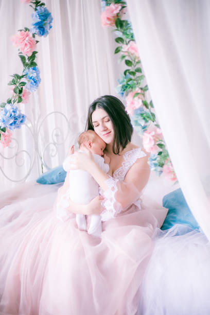 Young mother in a boudoir dress with a baby in her arms in spring flowers stock photo